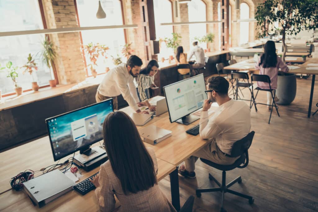 Équipe d'employés concentrés travaillant sur ordinateurs et collaborant dans un bureau open space lumineux avec fenêtres et plantes.