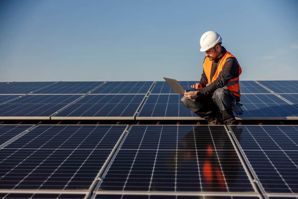 Ingénieur en gilet orange et casque blanc accroupi sur des panneaux solaires, utilisant un ordinateur portable sous ciel bleu. Surveillance énergie verte.