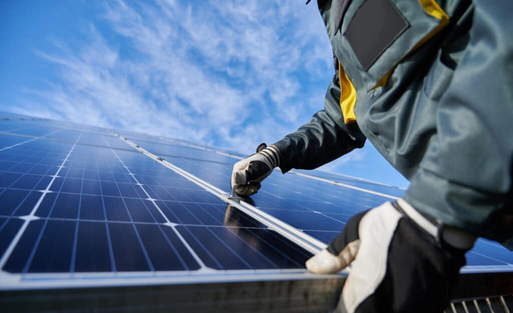 Main gantée d'un technicien fixant un panneau solaire bleu brillant. Ciel bleu clair avec quelques nuages en arrière-plan.