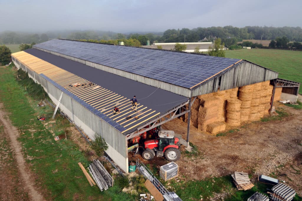 Ouvriers installant des panneaux solaires sur le toit d'une grande ferme avec foin et tracteur, entourée de champs brumeux.