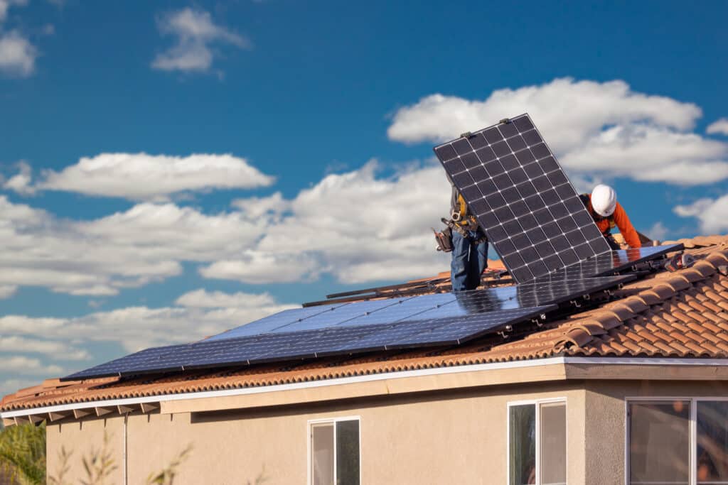 Deux techniciens installent des panneaux solaires photovoltaïques sur un toit de tuiles, sous un ciel bleu nuageux.