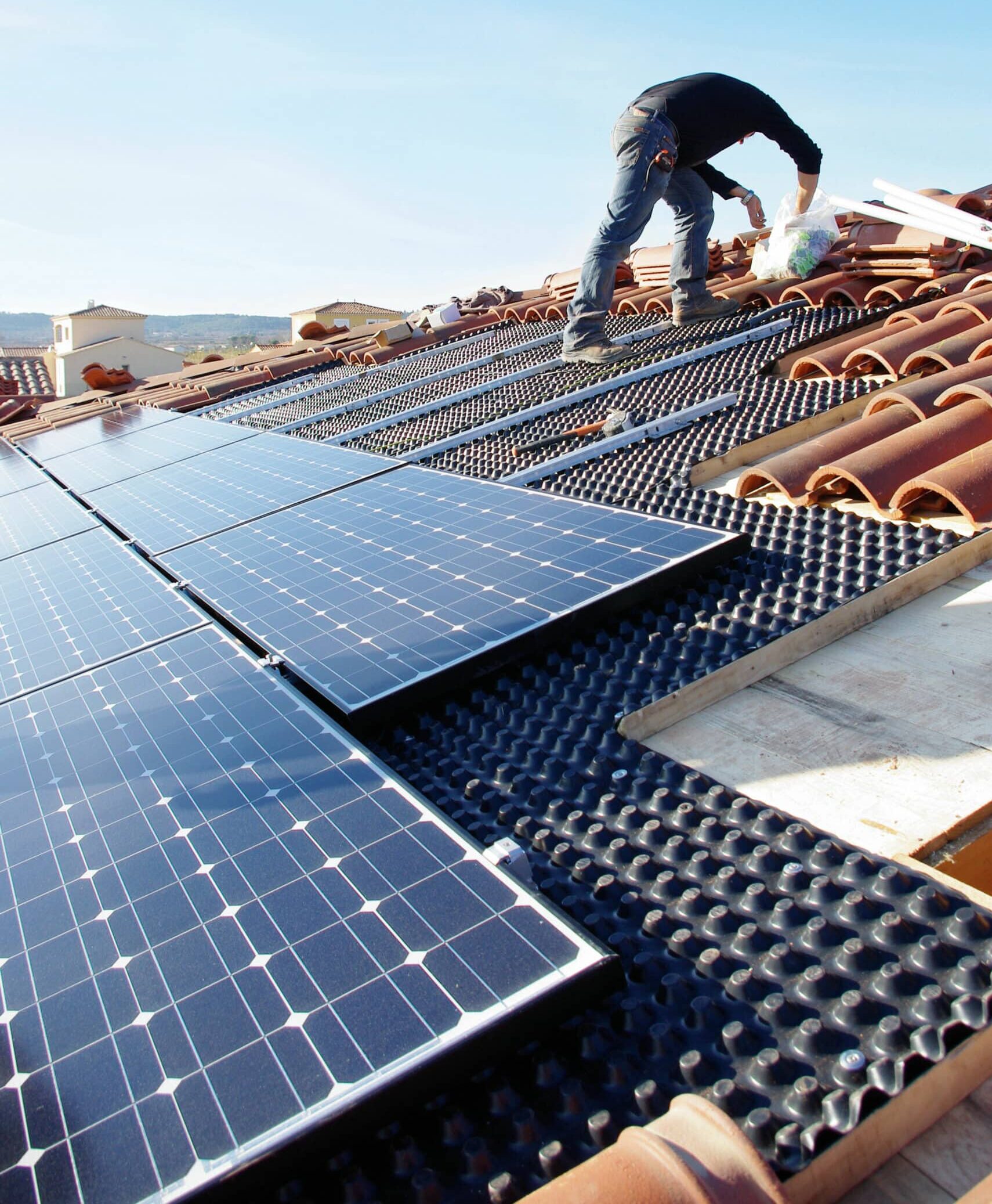 Installateur posant des panneaux solaires photovoltaïques sur un toit en tuiles, avec une sous-couche noire texturée. Ciel bleu.