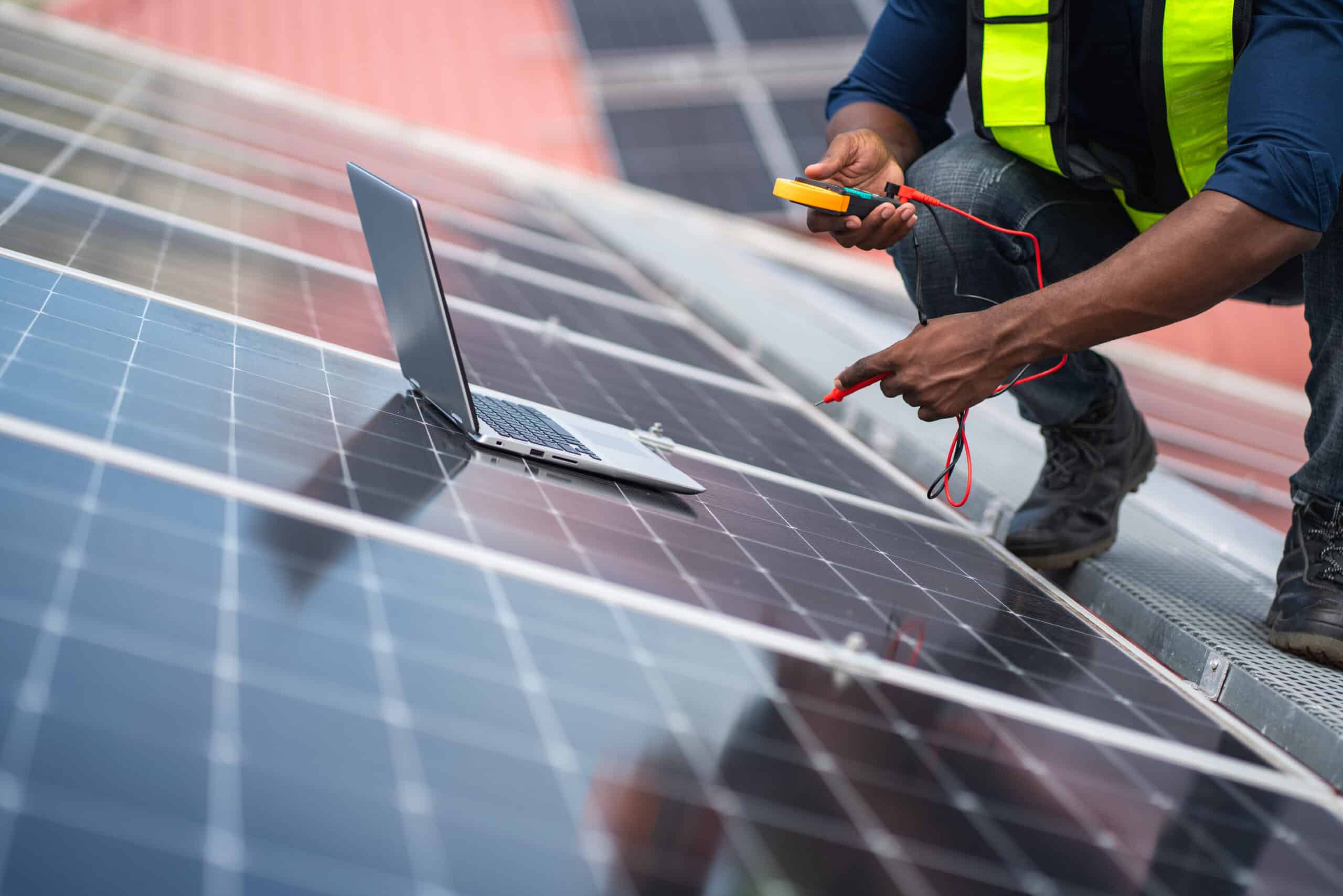 Technicien en gilet jaune sur un toit, testant des panneaux solaires avec un multimètre. Un ordinateur portable est posé à côté pour la surveillance.
