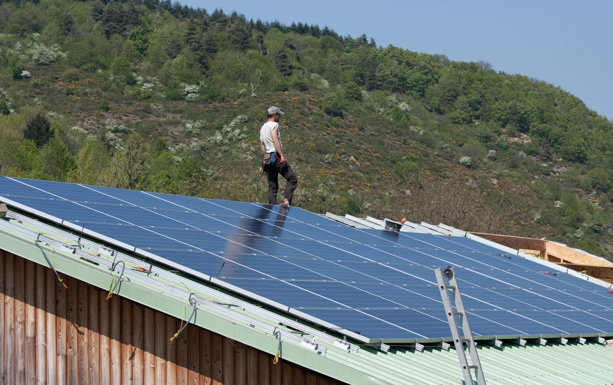 Homme en casquette installant des panneaux solaires bleus sur un toit vert. Montagne verdoyante en arrière-plan. Installation énergie solaire.