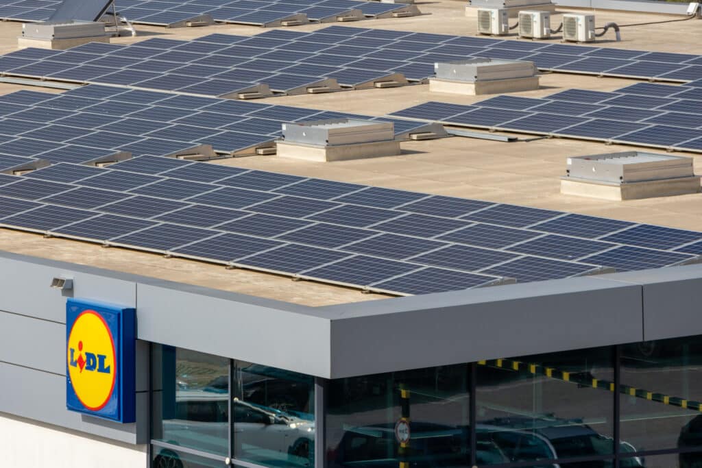 Toit d'un magasin Lidl couvert de nombreux panneaux solaires et unités de climatisation. Reflets de voitures dans les fenêtres.