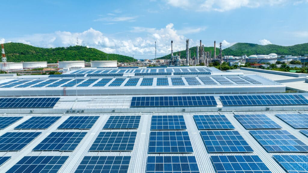 Toits d'usine couverts de panneaux solaires bleus, avec une raffinerie pétrolière et des collines verdoyantes sous un ciel nuageux.