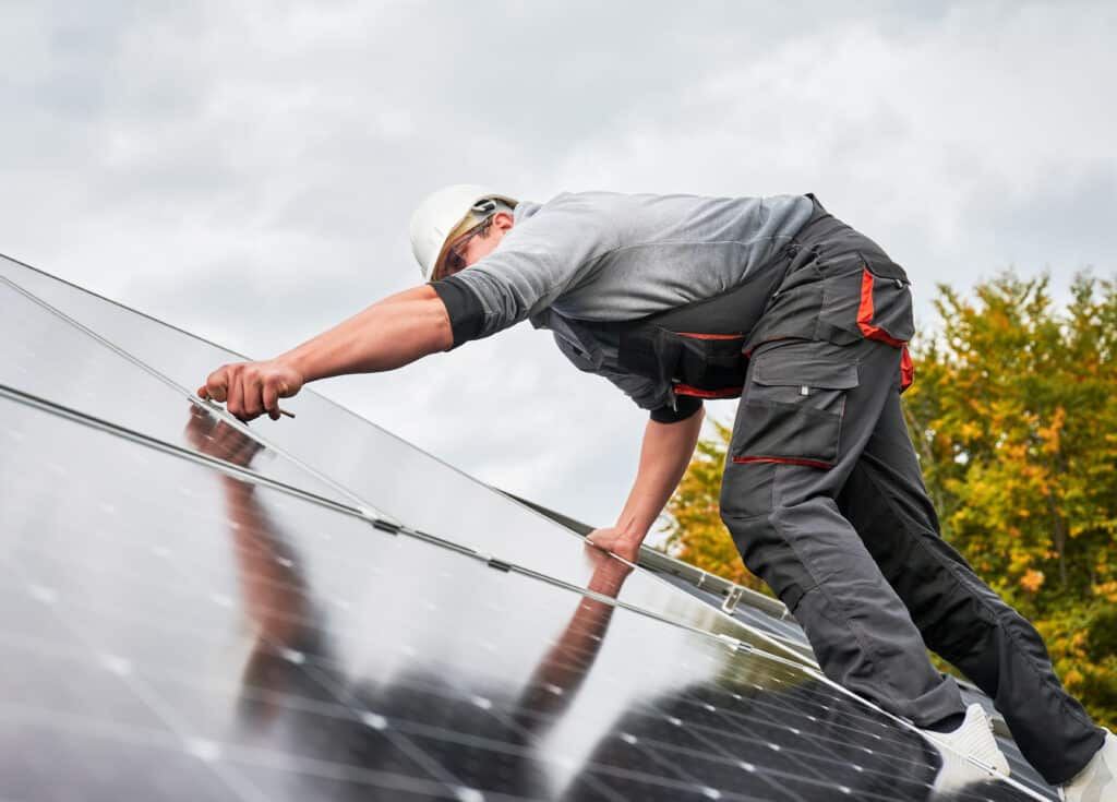 Technicien en casque et lunettes installant des panneaux solaires sur un toit. Reflet du ciel nuageux sur les modules.