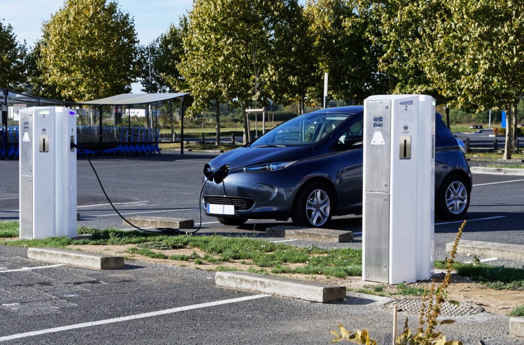 Voiture électrique grise en charge sur une borne de recharge 3kW dans un parking. Un câble noir connecte la voiture. Arbres verts en arrière-plan.