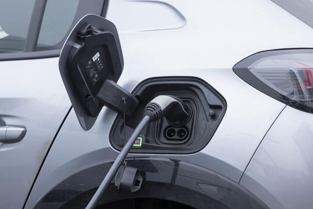 Voiture électrique grise en charge avec câble branché dans le port. Un voyant vert est actif et des symboles de recharge sont visibles.