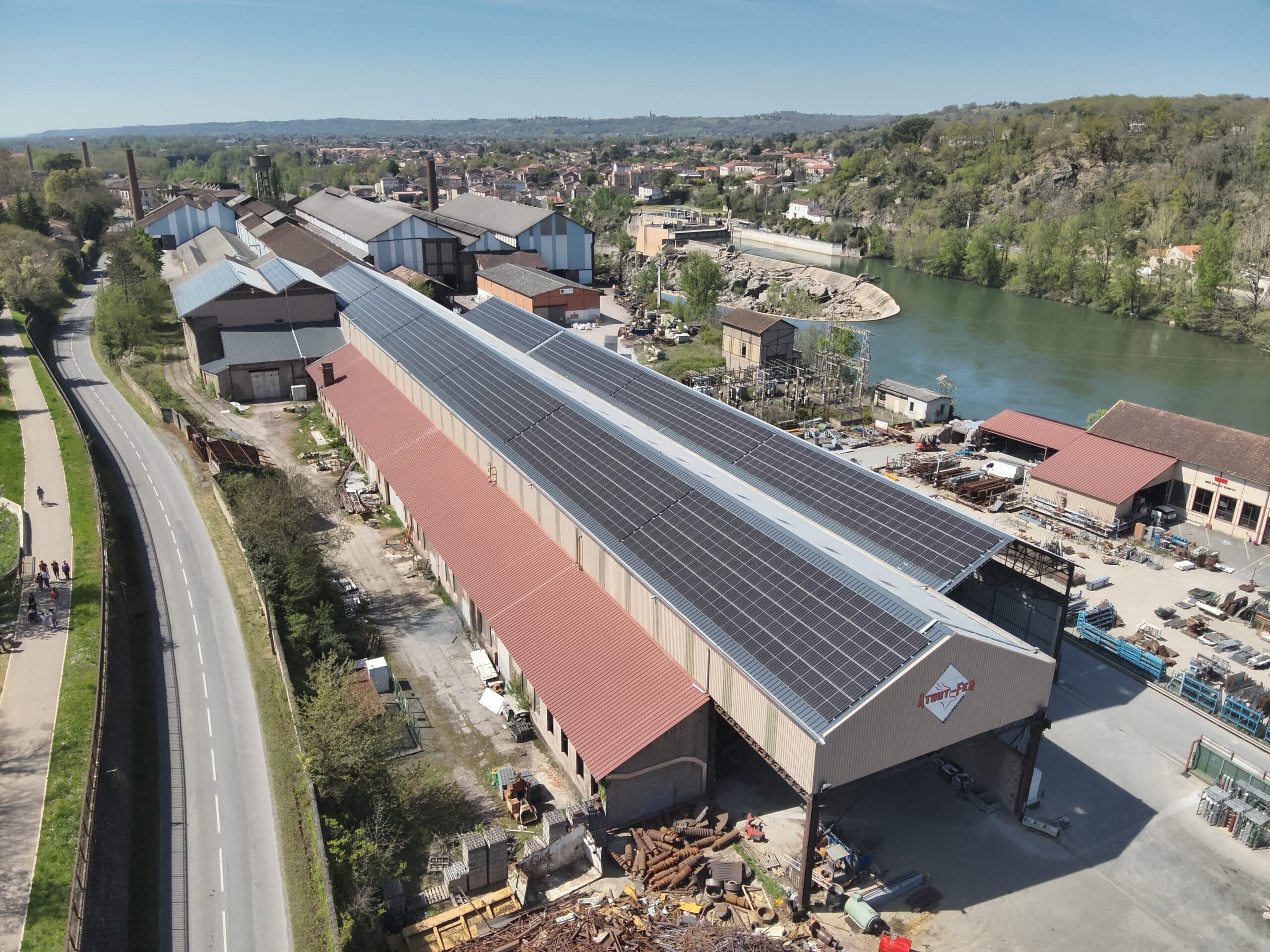 Vue aérienne d'un site industriel avec toits solaires, entre une route et une rivière, près d'une ville. Logo "A TOUT FER".
