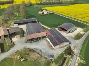Vue aérienne d'une ferme avec toits recouverts de panneaux solaires, entourée de champs de colza jaune et de cultures vertes.