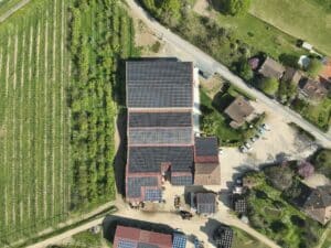 Vue aérienne d'un domaine agricole avec de vastes panneaux solaires sur les toits, à côté d'un vignoble en rangées et d'une zone arborée.