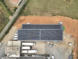 Vue aérienne d'un entrepôt industriel avec un vaste champ de panneaux solaires installés sur le toit, entouré de zones de terre et d'un parking avec des camions.
