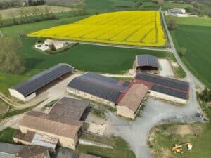 Vue aérienne d'une ferme avec toits photovoltaïques, champs de colza jaunes et prairies vertes. Un chariot élévateur JCB opère au premier plan.