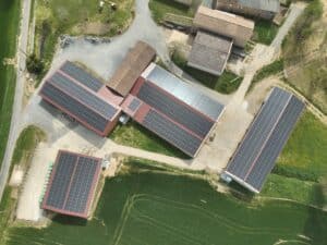 Vue aérienne ferme agricole toits couverts de panneaux solaires photovoltaïques au-dessus de champs verts et allées de gravier.