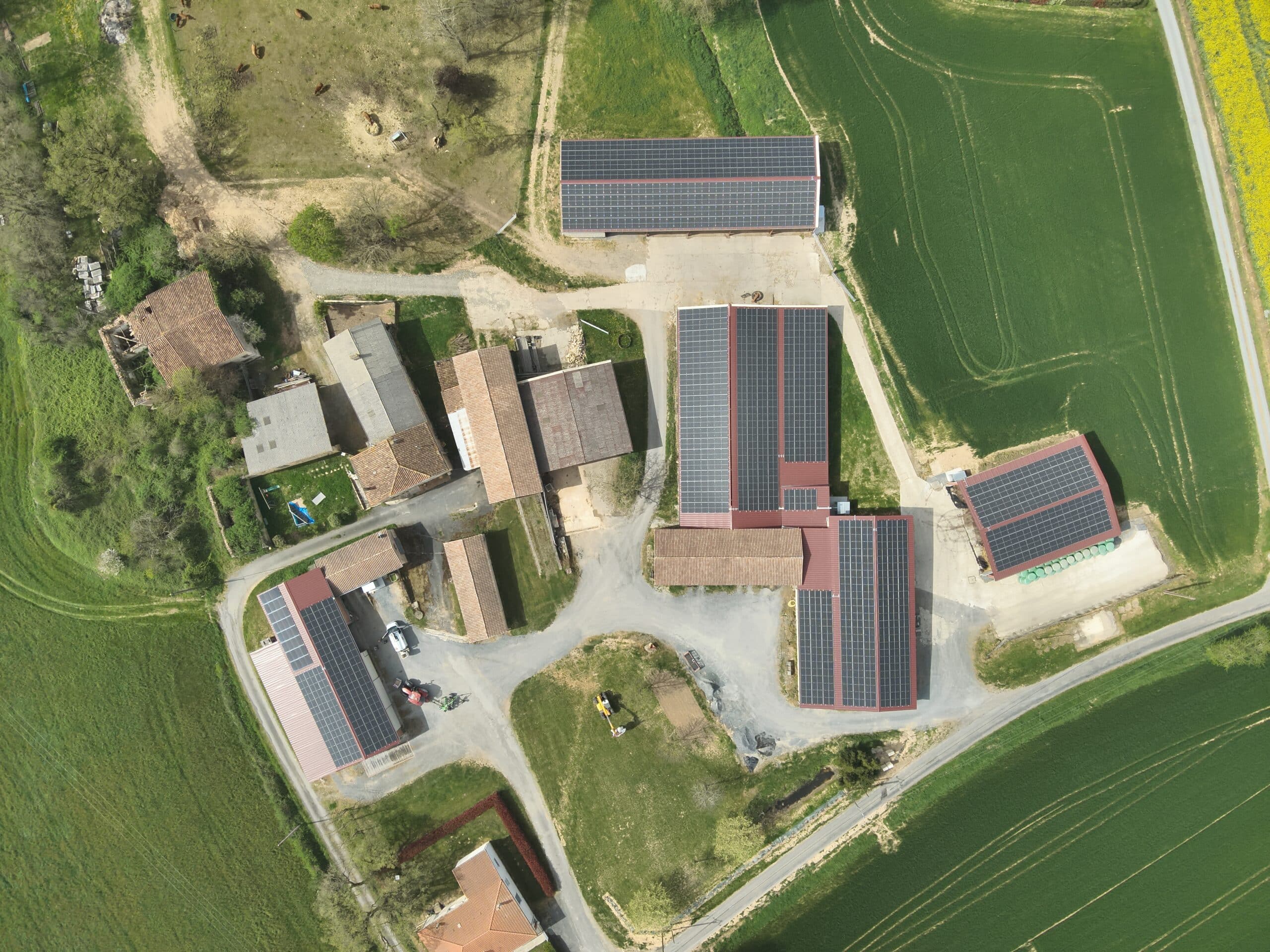 Vue aérienne d'une ferme rurale avec plusieurs bâtiments agricoles équipés de larges toits recouverts de panneaux solaires photovoltaïques, entourés de champs verts et de terre battue.