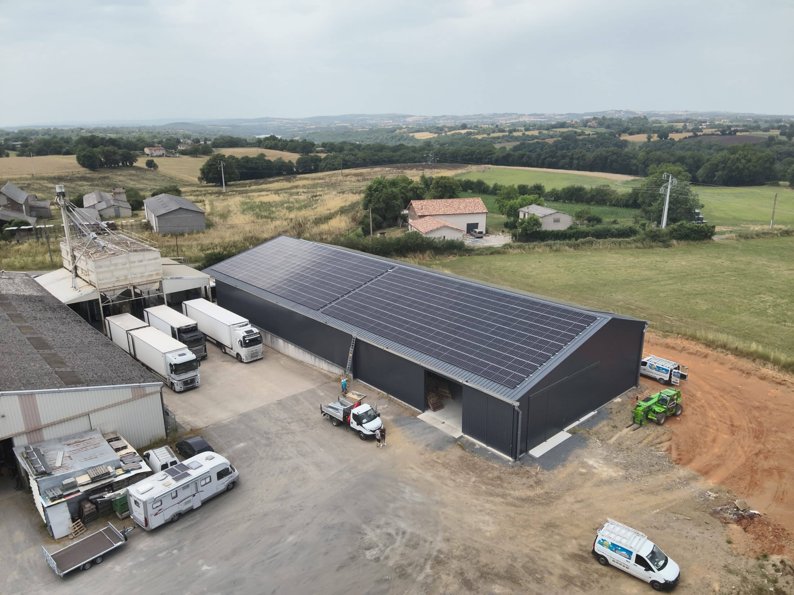 Vue aérienne d'un site industriel agricole avec un grand hangar équipé de panneaux solaires, camions et fourgons de service, dont un avec 'Spécialiste en énergie photovoltaïque'.