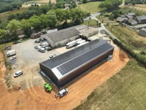 Vue aérienne d'un bâtiment industriel avec toit noir recouvert de panneaux solaires, entouré d'un chantier terreux et de verdure.