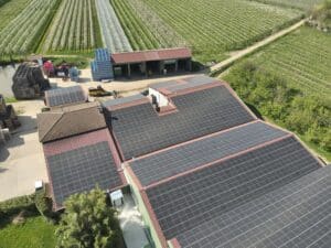Vue aérienne d'une exploitation agricole avec toits recouverts de panneaux solaires, entourée de vergers protégés par des filets blancs.
